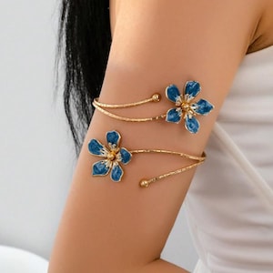May include: Gold arm cuff with two blue flower designs and gold centres. The floral accents are linked by delicate gold bands, creating an elegant accessory. This arm cuff is a stylish piece of jewellery, perfect for adding a touch of glamour.