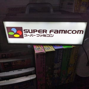 Op de afbeelding: Een wit verlicht bord met de tekst "SUPER FAMICOM" in zwarte letters en de Japanse tekst "スーパーファミコン" in kleinere zwarte letters. Het bord heeft een rood, blauw en geel logo in de linkerbovenhoek. Het bord is gemonteerd aan een muur boven een plank met verschillende videospelcartridges.