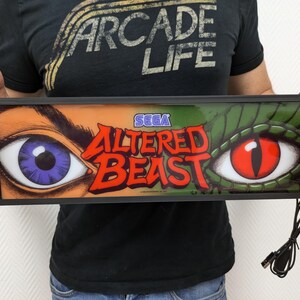 Può includere: Un cartello luminoso con cornice nera con un occhio verde e arancione e il testo "SEGA ALTERED BEAST".