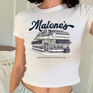 Può includere: Una t-shirt bianca a maniche corte con una grafica blu scuro raffigurante un diner. Il testo Malone's è scritto in corsivo sopra l'edificio e la scritta BEST BURGERS IN HASTINGS, MA è riportata sotto l'edificio al centro del petto.