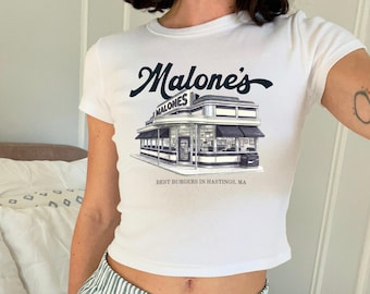 May include: A white short-sleeved t-shirt featuring a dark blue graphic of a diner building. The text reads Malone's in a script font above the building and BEST BURGERS IN HASTINGS, MA below the building. The design is centered on the chest.