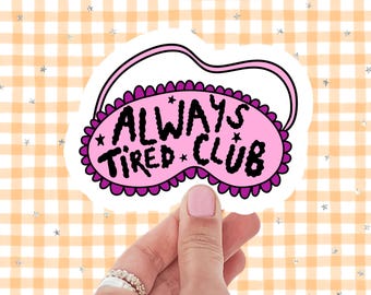 May include: A pink sleep mask sticker with the words "ALWAYS TIRED CLUB" in black. The mask has a scalloped edge and is decorated with small stars. The background is a checkered pattern in orange and white.