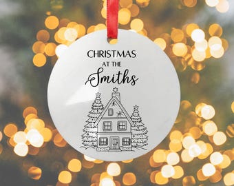 May include: White Christmas ornament with a red ribbon. The ornament features the text "CHRISTMAS AT THE Smiths" in black, above a detailed line drawing of a house with Christmas trees. The background is blurred.
