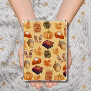 Pode incluir: Uma capa para tablet com um padrão de outono. O design inclui abóboras, fatias de torta, bolotas, livros, meias e folhas em tons de laranja, castanho e amarelo sobre um fundo bege. A capa é segurada por uma pessoa.