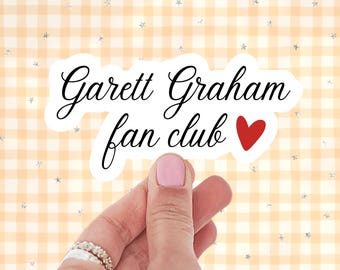 May include: A white die-cut sticker with a black cursive script that reads Garett Graham fan club, accompanied by a small red heart icon. The sticker features a clean white border and is presented against a light orange and white gingham background.