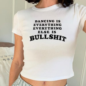 Könnte beinhalten: Ein weißes T-Shirt mit kurzen Ärmeln und schwarzem Text auf der Brust, der lautet: DANCING IS EVERYTHING EVERYTHING ELSE IS BULLSHIT. Das Kleidungsstück hat einen Rundhalsausschnitt und eine körpernahe Passform mit einem auffälligen Design.