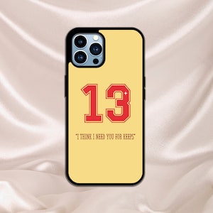 May include: A yellow iPhone case with a black border and a red number "13" in a white outline. The text "I think I need you for keeps" is printed below the number.