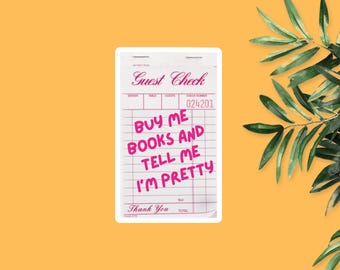 May include: A white guest check with pink text that reads "BUY ME BOOKS AND TELL ME I'M PRETTY." The check also has the words "Guest Check" and "Thank You" printed on it. The background is a solid orange colour.