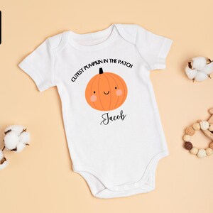 May include: White baby onesie with a smiling orange pumpkin graphic and the text "Cutest Pumpkin in the Patch" and the name "Jacob". The onesie is made of 100% cotton. Additional items include cotton buds and a wooden toy.