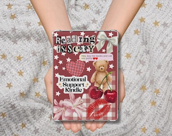 May include: A Kindle case with a burgundy background and white stars. The case features the text "Reading is Sexy" and "Emotional Support Kindle." Other graphics include a teddy bear, cherries, a bow, a heart, a flower, and a pair of red lips.