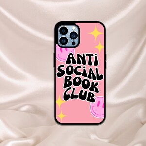 May include: Pink phone case with a black design that says "Anti Social Book Club". The design also includes two pink smiley faces with black outlines.