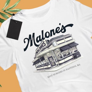 May include: A white t-shirt featuring a dark blue graphic of a retro diner building with the text "Malone's" above it and "BEST BURGERS IN HASTINGS, MA" below it. A black rectangular tag is attached to the collar with a thin black string.