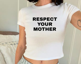 May include: A white short-sleeved t-shirt featuring the text "RESPECT YOUR MOTHER" printed in bold black capital letters across the chest. The garment has a crew neckline and a fitted silhouette, constructed from a plain white fabric.