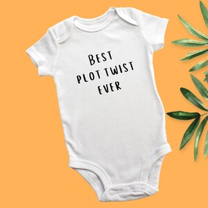 May include: White baby onesie with black text that reads "Best Plot Twist Ever".