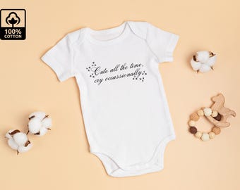 May include: White baby onesie with the text "Cute all the time, cry occasionally" in black script. The onesie is surrounded by cotton buds and a wooden toy horse on a peach-colored surface. The top left corner has a "100% cotton" logo.