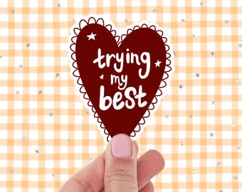 May include: A maroon heart-shaped sticker with a scalloped edge and the words "trying my best" in white script. The sticker is held against a background of orange and white gingham with silver star accents.