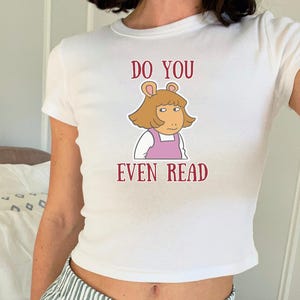 Puede incluir: Top corto blanco con una imagen de dibujos animados de D.W. Read de la serie de televisión Arthur. El texto granate "DO YOU EVEN READ" está impreso encima y debajo del personaje. La camiseta tiene mangas cortas y cuello redondo.