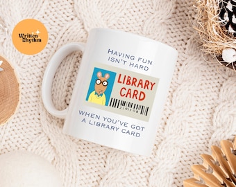 May include: White ceramic mug with a cartoon illustration of Arthur Read from the children's book series "Arthur." The mug has the text "Having fun isn't hard when you've got a library card" and a mock library card with the text "Library Card" and a barcode.