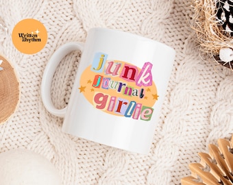 May include: A white ceramic mug featuring a colorful graphic design with the text Junk Journal Girlie in a collage-style font. The letters are set against a light orange oval background decorated with small yellow stars on a white surface.