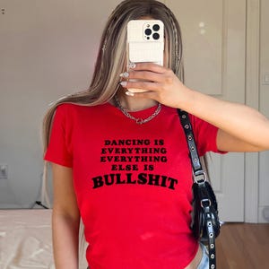Könnte beinhalten: Ein leuchtend rotes Kurzarm-T-Shirt mit schwarzem Textaufdruck auf der Vorderseite: DANCING IS EVERYTHING EVERYTHING ELSE IS BULLSHIT. Das T-Shirt hat einen Rundhalsausschnitt und eine körpernahe Passform aus einfarbigem Baumwollstoff.