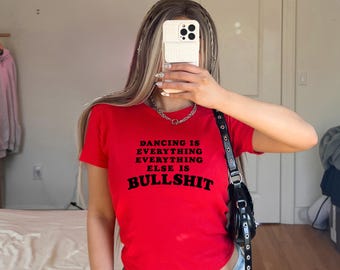 May include: A bright red short-sleeved t-shirt featuring black text printed on the front that reads: DANCING IS EVERYTHING EVERYTHING ELSE IS BULLSHIT. The shirt has a crew neckline and a fitted silhouette, made of a solid-colored cotton fabric.