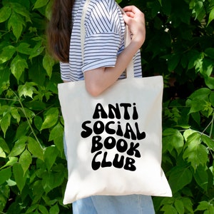May include: A white tote bag with black text that reads "Anti Social Book Club" in a retro font. The bag is being held by a person wearing a striped shirt.