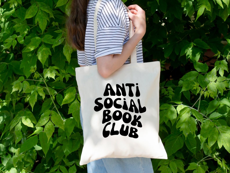 May include: A white tote bag with black text that reads "Anti Social Book Club" in a retro font. The bag is being held by a person wearing a striped shirt.