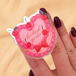Puede incluir: Una pegatina en forma de corazón con un diseño de pastel rosa, adornada con cerezas rojas y un lazo. La pegatina tiene las palabras "bookish girlie" en una fuente cursiva. La pegatina está sujeta por una mano con esmalte de uñas rojo oscuro.