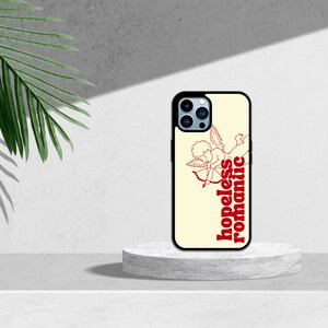 Puede incluir: Una funda de tel&eacute;fono blanca con borde negro. La funda tiene un dibujo de l&iacute;nea roja de un cupido con una flecha y el texto "hopeless romantic" en rojo.