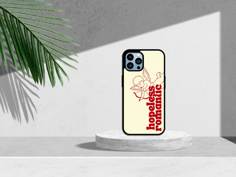 Puede incluir: Una funda de tel&eacute;fono blanca con borde negro. La funda tiene un dibujo de l&iacute;nea roja de un cupido con una flecha y el texto "hopeless romantic" en rojo.