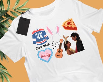 May include: A white t-shirt featuring a graphic print of a blue hockey jersey with the name Graham and number 44, pink bunny ears, a slice of cherry pie, a ukulele, musical notes, a heart with the text Wellsy, and a couple in costumes.