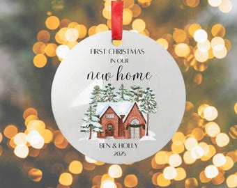 May include: A round Christmas ornament with a red ribbon. The ornament features a watercolor illustration of a house in a snowy forest, with the text "FIRST CHRISTMAS in our new home" and the names "BEN & HOLLY" and the year "2025".