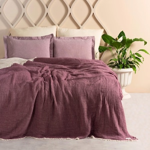 May include: A soft, mauve-colored throw blanket with a textured weave and a white fringe border. The blanket is draped over a bed with white and mauve pillows.