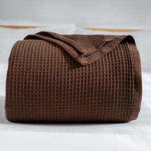 May include: A rolled-up, brown waffle-weave blanket. The blanket is a solid, dark brown color and has a textured, grid-like pattern. The blanket is folded over on itself, with a portion of the blanket draped over the top.
