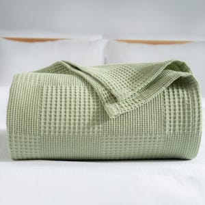 May include: A folded, light green waffle-weave blanket. The blanket is neatly arranged on a white surface, with a portion of the blanket folded over. The texture of the blanket is visible, with a grid-like pattern. The overall aesthetic is clean and simple.