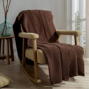 Pu&ograve; includere: Una coperta marrone a trama a nido d'ape drappeggiata su una sedia a dondolo in legno. La sedia ha seduta e braccioli fantasia. Un vaso di vetro con rami &egrave; su un tavolino di legno.