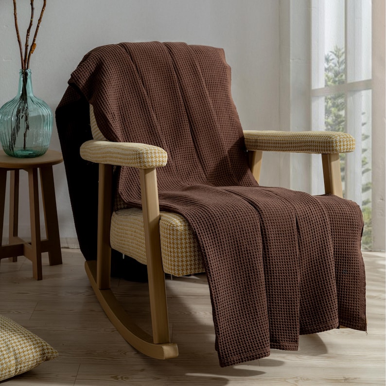 Pu&ograve; includere: Una coperta marrone a trama a nido d'ape drappeggiata su una sedia a dondolo in legno. La sedia ha seduta e braccioli fantasia. Un vaso di vetro con rami &egrave; su un tavolino di legno.