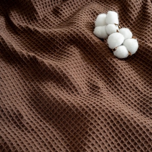 Pu&ograve; includere: Primo piano di un tessuto marrone a nido d'ape con una consistenza morbida. Un gruppo di batuffoli di cotone bianco poggia sul tessuto, aggiungendo un contrasto naturale alle calde tonalit&agrave; marroni. Il tessuto sembra essere una coperta o un asciugamano.
