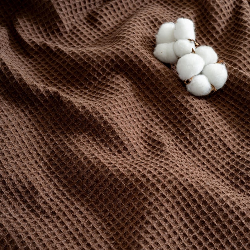 Pu&ograve; includere: Primo piano di un tessuto marrone a nido d'ape con una consistenza morbida. Un gruppo di batuffoli di cotone bianco poggia sul tessuto, aggiungendo un contrasto naturale alle calde tonalit&agrave; marroni. Il tessuto sembra essere una coperta o un asciugamano.