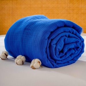May include: A rolled-up, vibrant blue blanket with a textured surface. Three cotton bolls are arranged in front of the blanket. The blanket is on a white surface, with a textured, yellow-orange background.