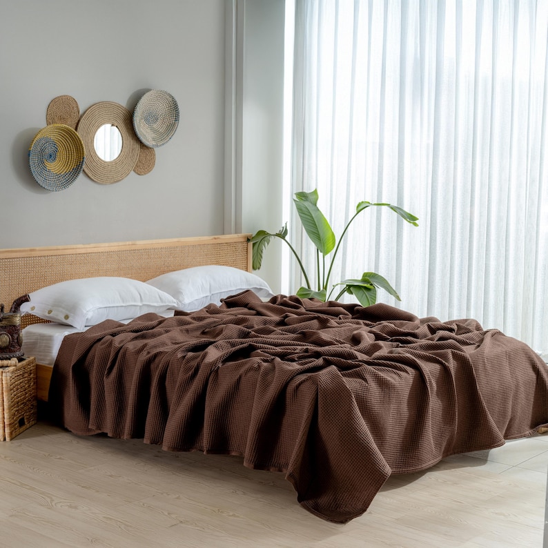 Pu&ograve; includere: Una coperta marrone a trama waffle copre un letto con cuscini bianchi. Il letto ha una testiera intrecciata. La stanza presenta una pianta e decorazioni murali. La coperta &egrave; di un colore caldo e neutro.