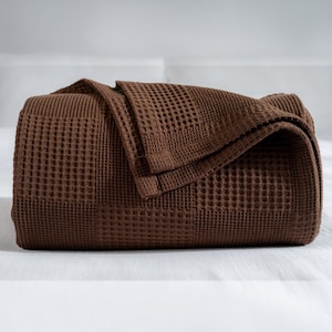 Peut inclure: Une couverture marron pliée en nid d'abeille. La couverture est d'un brun riche et chaud et présente un motif texturé en forme de grille. La couverture est pliée proprement, avec une partie drapée sur le dessus.