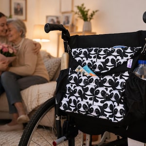 Può includere: Una borsa nera per sedia a rotelle con un motivo ripetuto di panda bianchi e neri. La borsa include un ampio scomparto con patta, una tasca frontale aperta e tasche laterali in rete che contengono una bottiglia d'acqua e altri oggetti.
