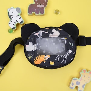 Può includere: Un marsupio per neonati in tessuto nero con una finestra in plastica trasparente e una cinghia nera regolabile. Il marsupio ha una divertente stampa di animali con una tigre, una zebra e altri animali.