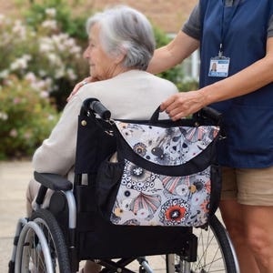 Op de afbeelding: Een zwarte opbergtas voor een rolstoel met een klep voorzien van cirkelvormige bloemmotieven in oranje, zwart en wit. De tas is uitgerust met een handvat aan de bovenzijde, zijvakken en verstelbare riemen voor bevestiging aan het frame.