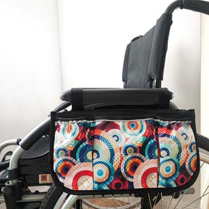 Puede incluir: Bolsa para silla de ruedas con un vibrante patrón de círculos superpuestos en tonos rojos, azules, naranjas y turquesas. La bolsa está sujeta a la parte delantera de una silla de ruedas negra, ofreciendo almacenamiento. Tiene cremallera y ribete negros.