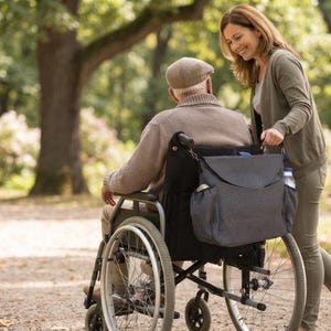 Peut inclure: Un sac de rangement gris foncé pour fauteuil roulant avec une fermeture à rabat et des poches latérales, fixé à l'arrière d'un fauteuil roulant. Le sac présente une finition en tissu texturé et une bouteille d'eau est visible dans une poche.