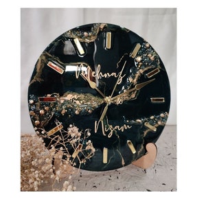 Op de afbeelding: Een ronde, zwarte en gouden harsklok met een marmer effect en gouden glitter. De wijzerplaat van de klok heeft gouden wijzers en gouden Romeinse cijfers. De klok is gepersonaliseerd met de namen "Mehnaz & Nizam".