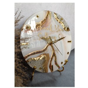 Könnte beinhalten: Weiße und goldene Harz-Uhr mit marmormusterartigem Design und goldenen Glitzerakzenten. Das Zifferblatt der Uhr hat römische Ziffern und die Worte "Thank You Sister" in goldener Schrift. Die Uhr steht auf einem goldenen Metallständer.