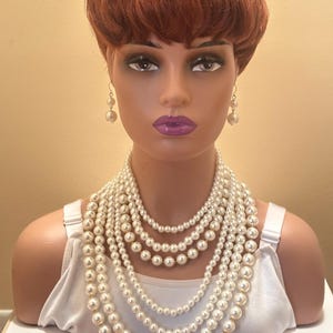May include: A mannequin head with a red wig and pearl jewellery. The jewellery includes a set of pearl earrings and multiple strands of white faux pearl necklaces. The mannequin is wearing a white vest top.